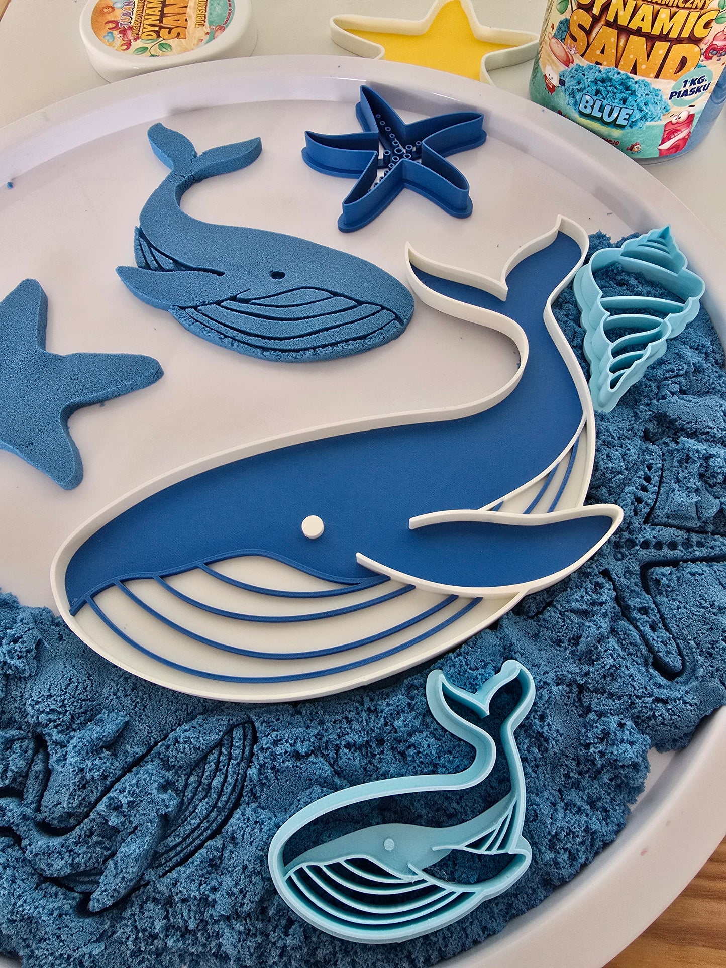 Blauwe walvisvormen en oceaansnijders op een witte speeltray gevuld met blauw kinetisch zand, inclusief een grote 3D-geprinte walvis, zeester, schelp en afdrukken in het zand.