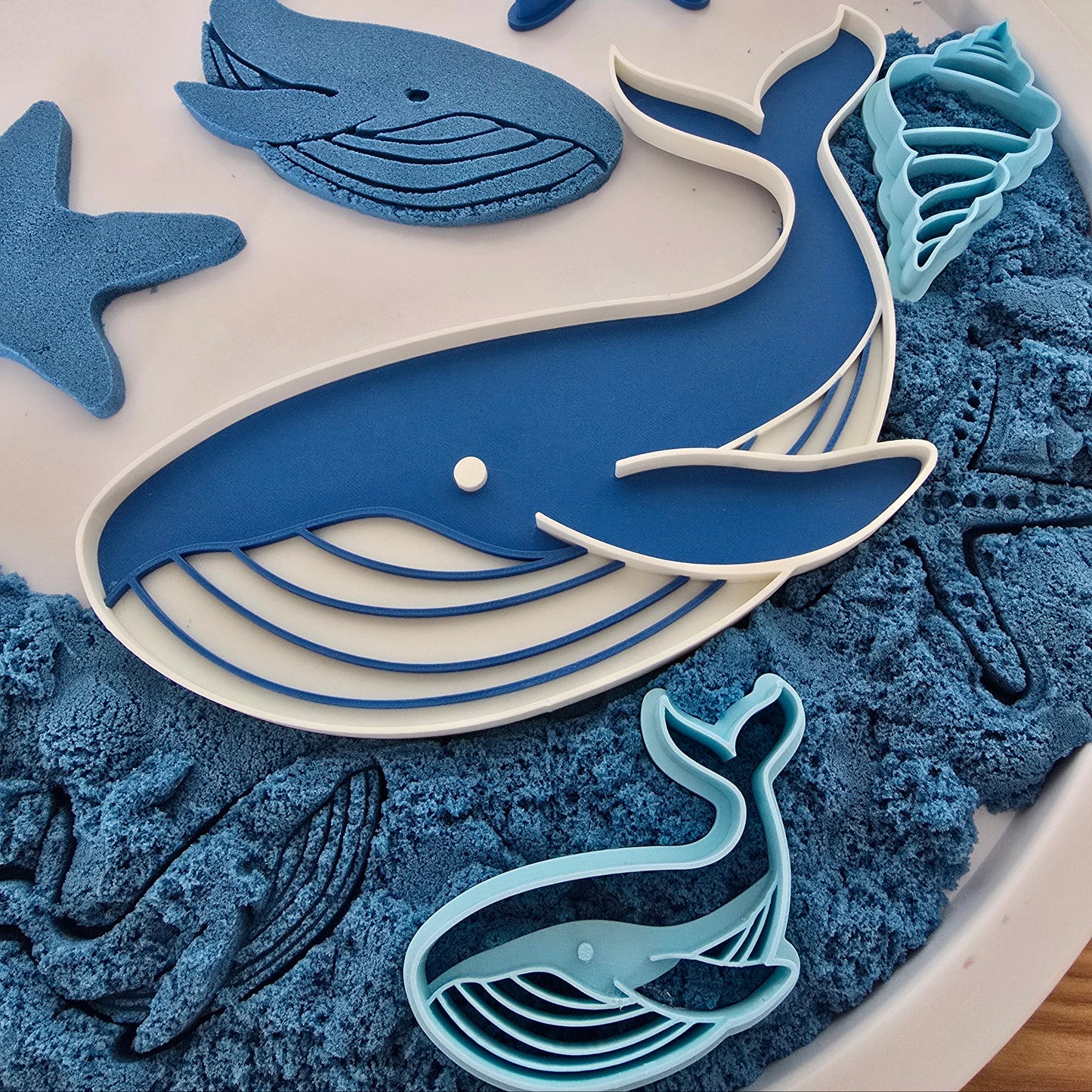 Blauwe walvisvormen en oceaansnijders op een witte speeltray gevuld met blauw kinetisch zand, inclusief een grote 3D-geprinte walvis, zeester, schelp en afdrukken in het zand.