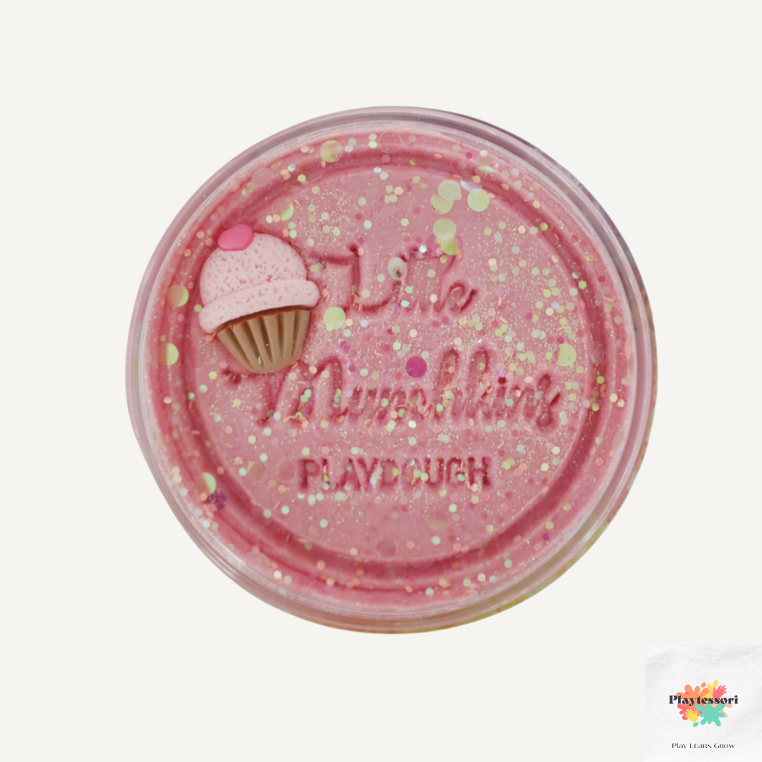 Een close-up van een potje roze speeldeeg met het logo van 'Little Munchkins PLAYDOUGH' in reliëf in het deksel. Het deeg is overvloedig bestrooid met glitters. Aan de linkerkant van het deksel is een kleine, roze en bruine cupcake-versiering bevestigd.