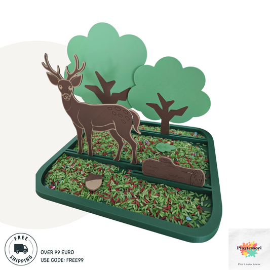A forest-themed sensory play tray designed for children, featuring a 3D scene with a brown deer, three green trees, a wooden log, an acorn, and colorful rice representing grass and forest floor. The tray is sectioned and bordered in green plastic.