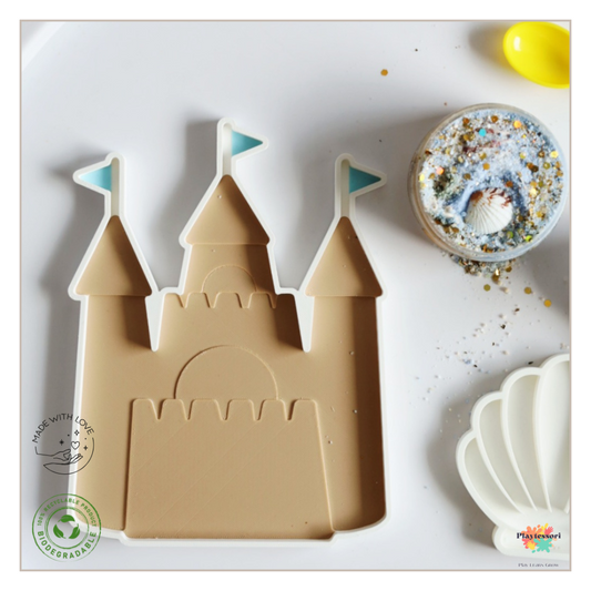 An overhead shot of a beige, 3D-printed sandcastle mold with three towers and two light blue flags on a white background. To the right is a small, clear container filled with blue, white, and tan botanical fizz, topped with a small seashell and gold glitter.