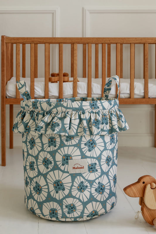 Speelgoedmand met denim leeuwenprint en volant in een babykamer naast een houten ledikant