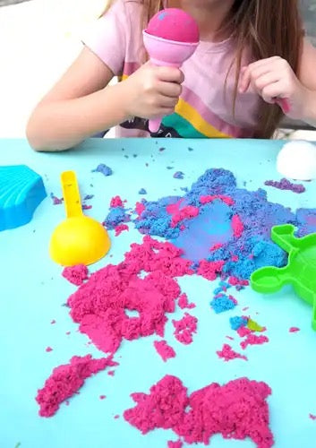 Een meisje speelt aan tafel met roze en paarse dynamisch zand. Ze houdt een roze ijsvorm vast en is omringd door kleurrijke zandvormpjes, waaronder een groene schildpad en een gele schep.