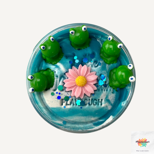  Een close-up van een potje speeldeeg met een vijverthema. Het deeg is blauw en wit gemarmerd en bevat vijf groene, plastic kikkertjes, een roze bloem met een gele kern, en blauwe glitters.