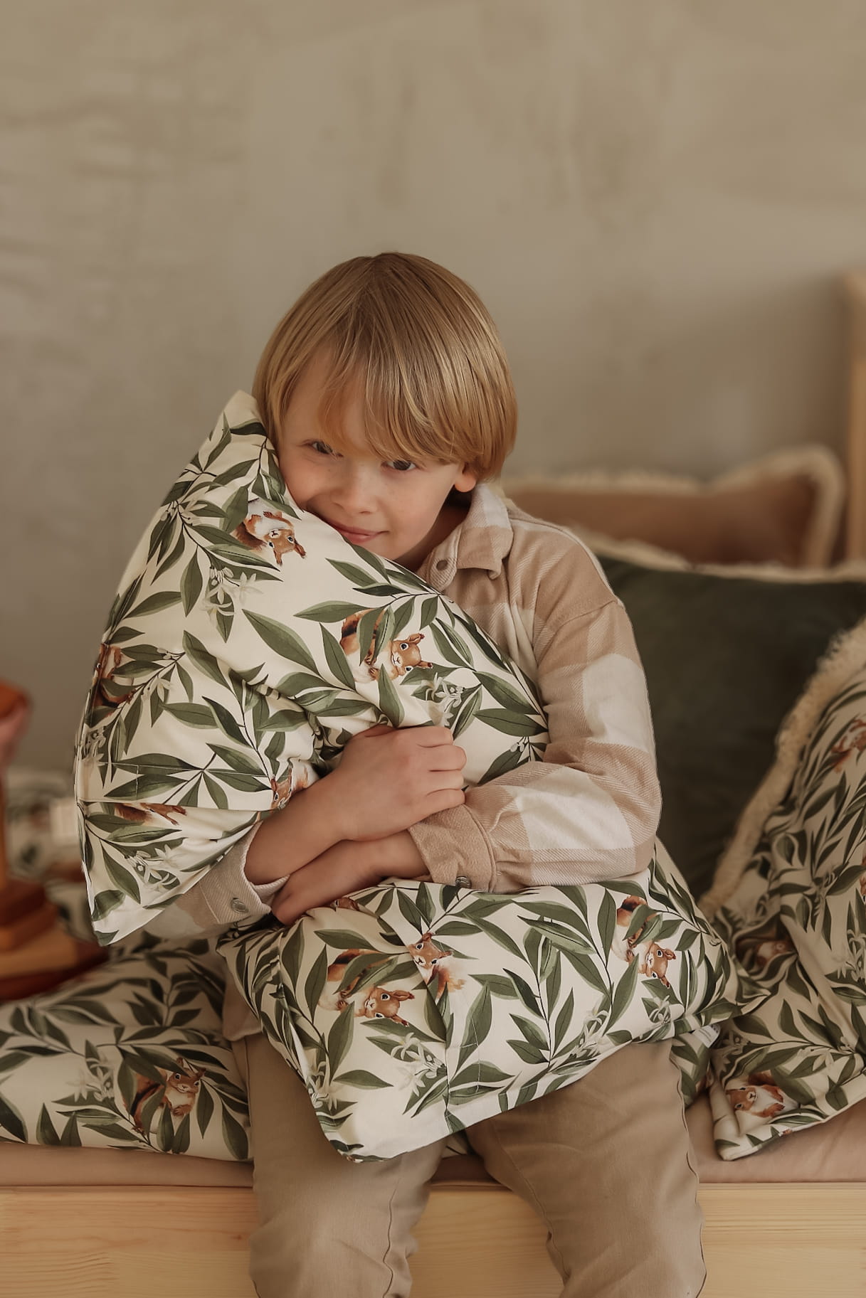 Lachend kind zit op een houten bed en knuffelt een kussen met bladeren- en eekhoornprint. De sfeer is warm en huiselijk, met natuurlijke kleuren en zachte materialen op de achtergrond.