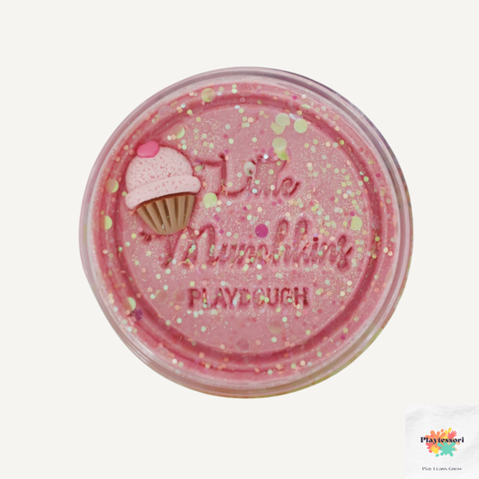 Een close-up van een potje roze speeldeeg met het logo van 'Little Munchkins PLAYDOUGH' in reliëf in het deksel. Het deeg is overvloedig bestrooid met glitters. Aan de linkerkant van het deksel is een kleine, roze en bruine cupcake-versiering bevestigd.